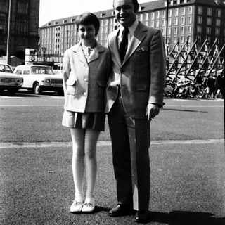 Two people posing together in formal attire, 1950s-60s urban setting.