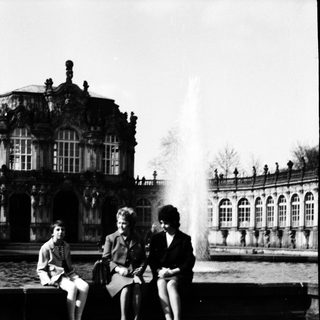Three people sitting by a fountain in front of historic palace.