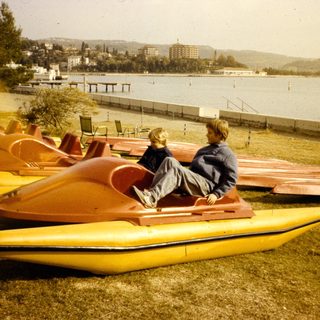 Two people sitting on pedal boats by a lake.