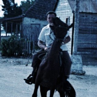 Man riding horse on coastal road near rustic buildings.