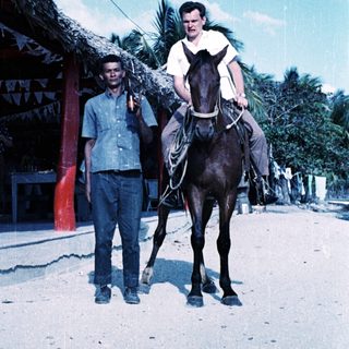 Two men: one riding horse, one standing beside. Tropical setting.