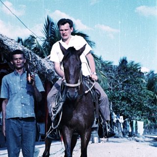 Two men: one riding horse, one standing beside. Tropical setting.