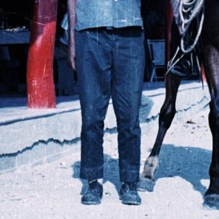 Two men: one riding horse, one standing beside. Tropical setting.
