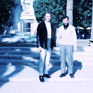 Two men posing outdoors near historic building with palm trees.