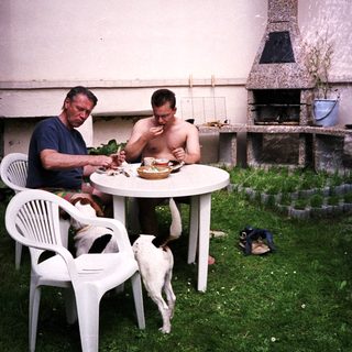 Two men eating outdoors with a dog nearby.