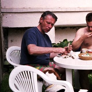 Two men eating outdoors with a dog nearby.