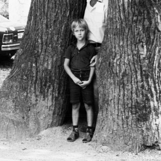 Two people posing beside a large tree. Classic cars parked nearby.