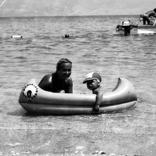Two people in inflatable raft near water, with motorboats and swimmers in background.