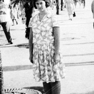 Vintage seaside promenade with palm trees and mid-20th-century attire.