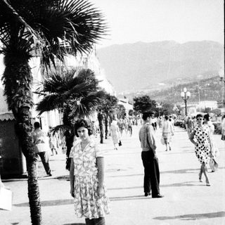 Vintage seaside promenade with palm trees and mid-20th-century attire.