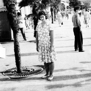 Vintage seaside promenade with palm trees and mid-20th-century attire.