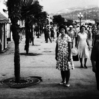 Mid-20th-century seaside promenade with palm trees and people.