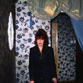 A woman in a dark blazer stands in a dimly lit, vintage floral-patterned room.