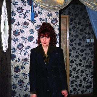 A woman in a dark blazer stands in a dimly lit, vintage floral-patterned room.