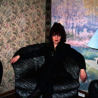 Person wearing large black coat indoors near floral wallpaper.