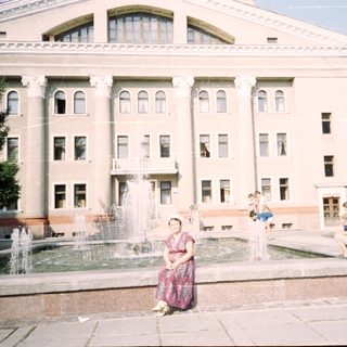 A woman sits near a fountain in front of a large building.