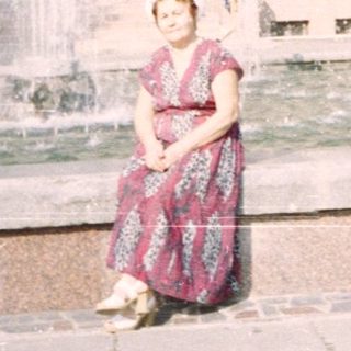 A woman sits near a fountain in front of a large building.