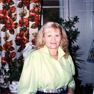 Woman in green blouse posing indoors near floral curtain.