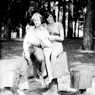 Two people posing together outdoors on tree stumps.