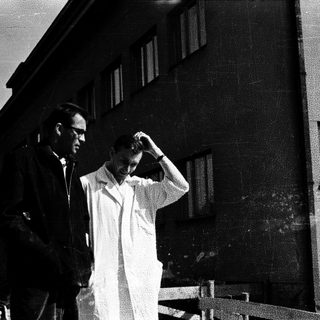 Here's a brief summary of the image: Two men stand outside a building, appearing contemplative.