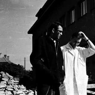 Here's a brief summary of the image: Two men stand outside a building, appearing contemplative.