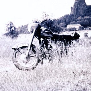 Motorcycle sits in overgrown field with ruins in background.
