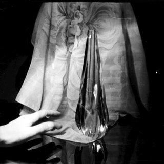 Here's a brief summary of the image: A hand near a glass vase, dark background.