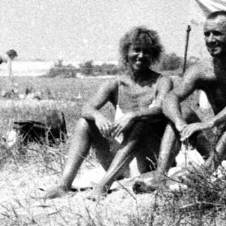 Two men seated in tall grass near a body of water.