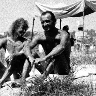 Two men sit in a grassy area, people in background.