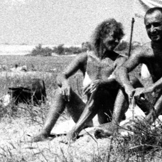 Two men sit in a grassy area, people in background.
