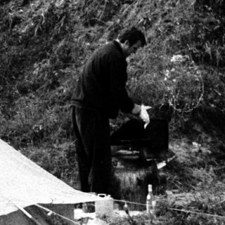 Man stands near a tent in a grassy, wooded area.