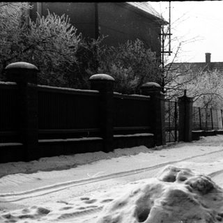 Snow-covered road with fence, houses, and trees visible.
