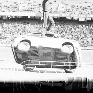 Man stands atop overturned car in stadium crowd.