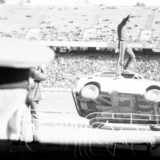 Man stands atop overturned car in stadium crowd.