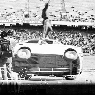 Man stands atop overturned car in stadium during event.