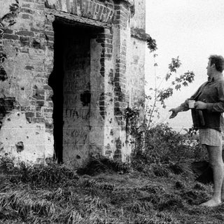 Man stands near ruins, holding a cup by the water.