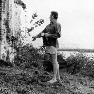 Man stands near ruins, holding a cup by the water.