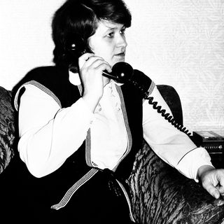 Here's a brief summary of the image: Woman on a vintage telephone, seated on a sofa.
