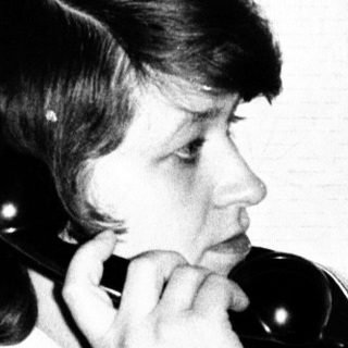 Woman seated, holding a vintage telephone to her ear.
