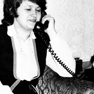 Woman sits, holding a telephone, in a vintage setting.
