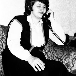 Woman sits, holding a telephone, in a vintage setting.
