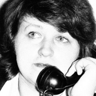 Here's a brief summary of the image: Woman seated, holding a vintage telephone to her ear.