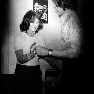 Man and woman standing, one smoking, in a dimly lit room.