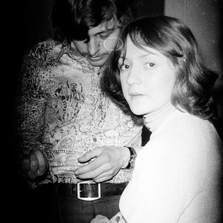 Here's a brief summary of the image: A couple stands close together in a dimly lit room.