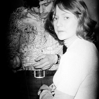 Here's a brief summary of the image: A couple stands close together in a dimly lit room.