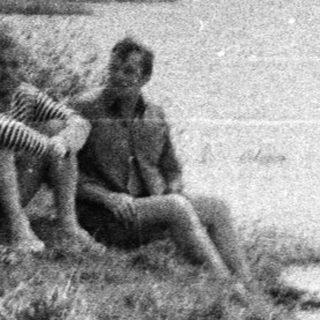 Two people sit near a ruin by a lake.