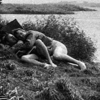 Two figures rest near a ruin by a body of water.