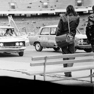 Cars drive past an empty stadium; people watch.