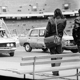 Cars drive past an empty stadium; people watch.