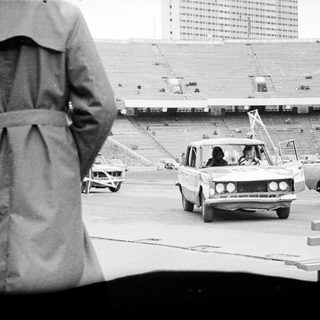 Cars drive past an empty stadium; people watch.